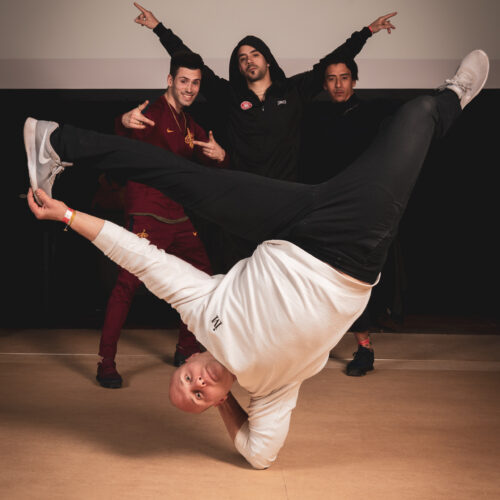 Quatre personnes posent pour une photo, avec un danseur effectuant une figure acrobatique au premier plan. Les autres personnes se tiennent debout derrière lui, faisant des gestes de victoire. Le fond présente le logo 'K-Rément Hip Hop'