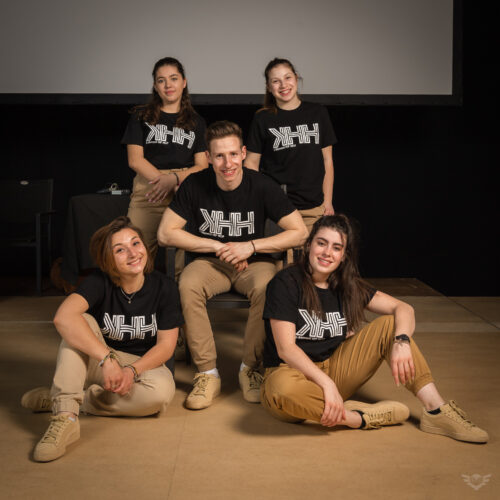 Un groupe de cinq personnes posant pour une photo, toutes portant des t-shirts noirs avec le logo KHH, sur fond sombre avec des lumières douces.