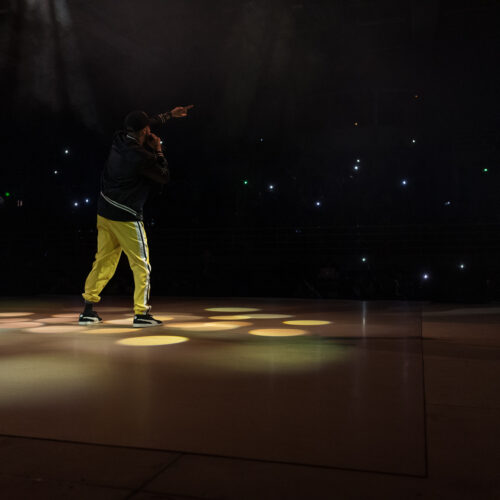 Un artiste sur scène pointe vers le public, entouré de lumières de scène et de téléphones allumés dans le public.