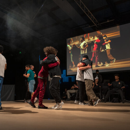 Des danseurs se félicitent en s'embrassant ou se serrant dans les bras, avec un écran montrant la scène en arrière-plan.