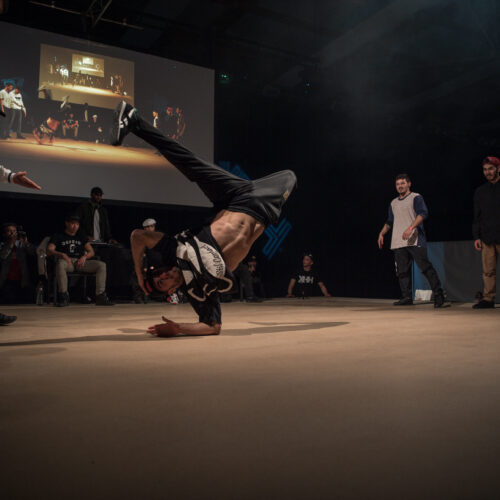 Un danseur effectue un mouvement de breakdance au sol, tandis que des spectateurs l'encouragent autour de lui. La scène est éclairée par des lumières vives, et un grand écran à l'arrière projette une image du même danseur.