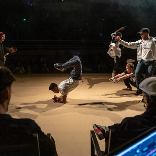 Un danseur effectue un mouvement de breakdance au sol, tandis que d'autres danseurs en arrière-plan posent des gestes expressifs. Des spectateurs et un DJ sont visibles sur scène.