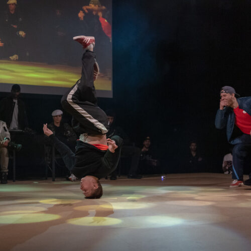 Un danseur exécute un mouvement de breakdance à l'envers, tandis que d'autres danseurs réagissent avec enthousiasme et un écran en arrière-plan projette une image de la performance.