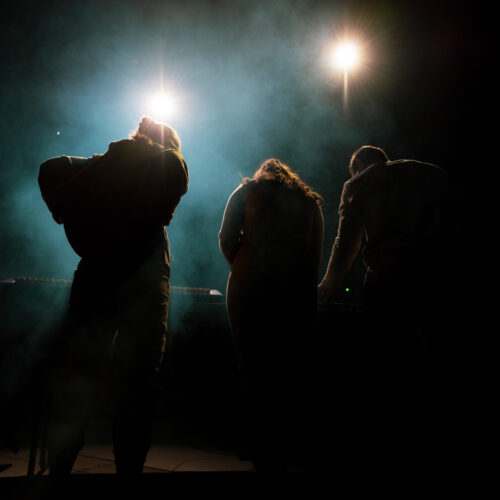 Trois personnes sont vues de dos, manipulant un équipement musical sous des projecteurs lumineux, créant un effet de contre-jour avec de la fumée.