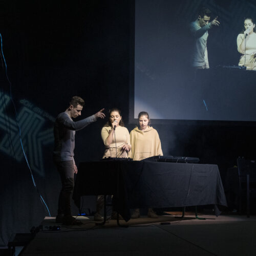 Un animateur et deux artistes en tenues beiges interagissent sur scène, l'un avec un micro et les autres près d'un contrôleur musical, tandis qu'une projection de leur performance est affichée en arrière-plan.