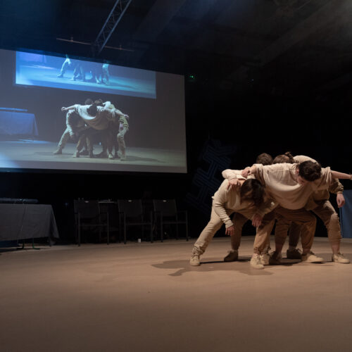 Un groupe de danseurs effectue une pose collective sur scène, capturée en projection derrière eux, tandis qu'une personne manipule la musique à la table de mixage.