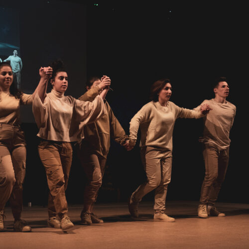Un groupe de danseurs vêtus de tenues beiges se tiennent par la main en exécutant une chorégraphie sur scène, avec une projection de leur performance en arrière-plan.
