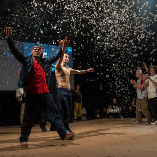 Des danseurs sur scène saluent le public sous une pluie de confettis, tandis qu'un autre danseur lève les bras en signe de célébration. L'arrière-plan montre des spectateurs applaudissant.