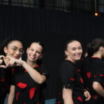 Trois jeunes femmes souriantes, portant des t-shirts noirs avec des motifs rouges, font un geste de cœur avec leurs mains sur scène.