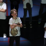 Des enfants portant des t-shirts blancs et des horloges suspendues autour du cou, les bras croisés, sur scène sous des projecteurs.