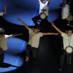 Des enfants portant des t-shirts blancs et des chaînes avec de grandes horloges exécutent une chorégraphie, en position accroupie, sous des projecteurs