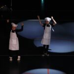 Deux enfants portant des tabliers et des toques de chef effectuent une chorégraphie sur scène sous des projecteurs.