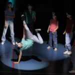 Un groupe de danseurs portant des masques colorés observe un membre du groupe exécutant un mouvement de breakdance sur scène, éclairé par des projecteurs.