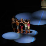 Cinq enfants, pieds nus, forment une chaîne et dansent sous des projecteurs qui éclairent le sol de manière circulaire.