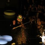 Un animateur frappe une piñata colorée avec un bâton, faisant tomber des bonbons et des confettis, tandis qu'un groupe d'enfants regarde avec enthousiasme