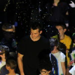 Un animateur en t-shirt noir sourit parmi un groupe d'enfants, avec des confettis flottant autour d'eux sur scène