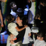 Une animatrice souriante porte une petite fille sur scène, entourée d'autres enfants vêtus de t-shirts blancs, avec des confettis colorés dans leurs cheveux.