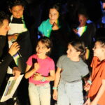 Un animateur s'adresse à un groupe d'enfants sur scène, tandis que quatre enfants sourient et écoutent attentivement, certains portant des t-shirts colorés.