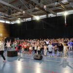Un grand groupe d'enfants dansant en synchronisation sous la direction d'un instructeur, avec des mouvements de bras levés et un décor de scène