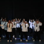 Groupe de danseurs en chemises blanches et cravates noires, exécutant une chorégraphie synchronisée sur scène.