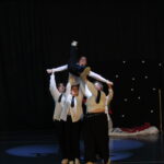 Un groupe de danseurs en chemises blanches et cravates noires réalise une figure acrobatique impressionnante, soulevant une danseuse au-dessus de leurs têtes. La scène est éclairée par des projecteurs, mettant en valeur leur synchronisation et leur force. L'arrière-plan sombre contraste avec la tenue des participants et les motifs décoratifs.