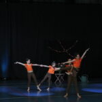 Trois jeunes danseuses vêtues de hauts orange vif et de leggings marron exécutent une chorégraphie gracieuse sur une scène éclairée. Une ambiance florale et festive est créée en arrière-plan avec des guirlandes lumineuses et des décorations florales.