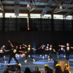 Groupe de danseurs en tenue noire avec des t-shirts ornés de motifs de crâne, réalisant une chorégraphie synchronisée dans un gymnase.