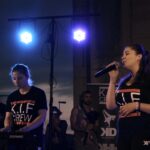 Une chanteuse tient un micro sur scène, tandis qu'une autre personne joue du clavier. Les deux portent des t-shirts noirs avec le logo 'K.I.F CREW', sous des projecteurs bleus.