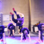 Un danseur effectue un saut acrobatique pendant une performance sur scène, entouré d'autres danseurs en position de préparation, sous des projecteurs lumineux.