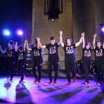 Des danseurs du groupe K.I.F CREW et K.I.F KIDZ se tiennent par la main sur scène, saluant le public sous des projecteurs lumineux.