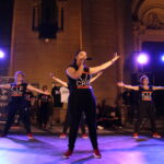 Un groupe de danseurs sur scène, vêtus de t-shirts noirs avec le logo 'K.I.F CREW', effectue une chorégraphie sous des projecteurs, avec un membre du groupe tenant un micro et en train de chanter.
